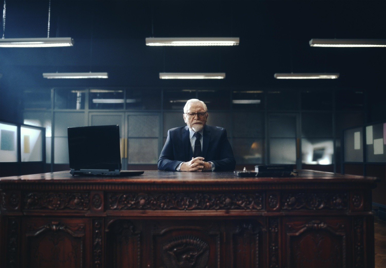 Actor Brian Cox seated at an imposing wooden desk in a corporate office 