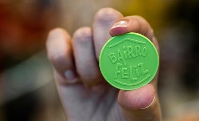 Fingers holding a green plastic 'Bairro Feliz' token 