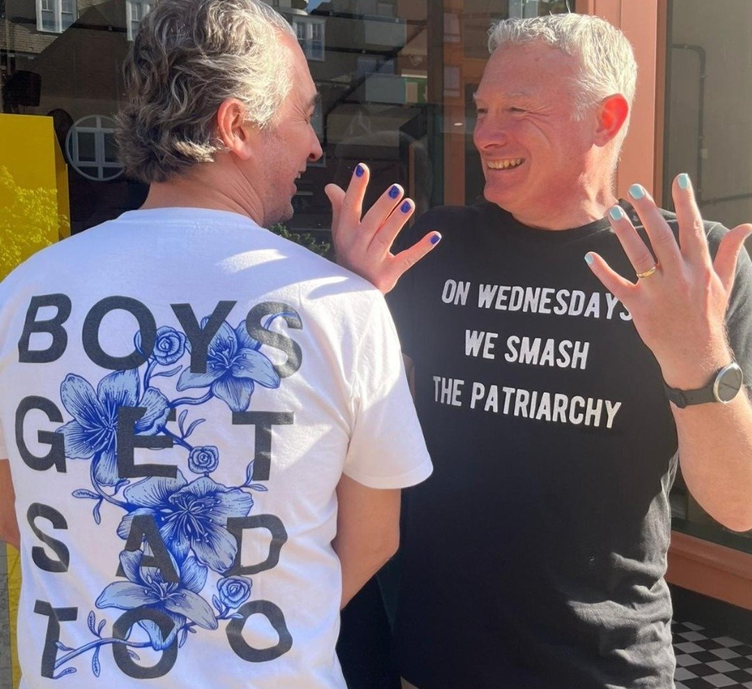 Two middle-aged men, one wearing nail polish, the other a t-shirt reading 'Boys get sad too' 