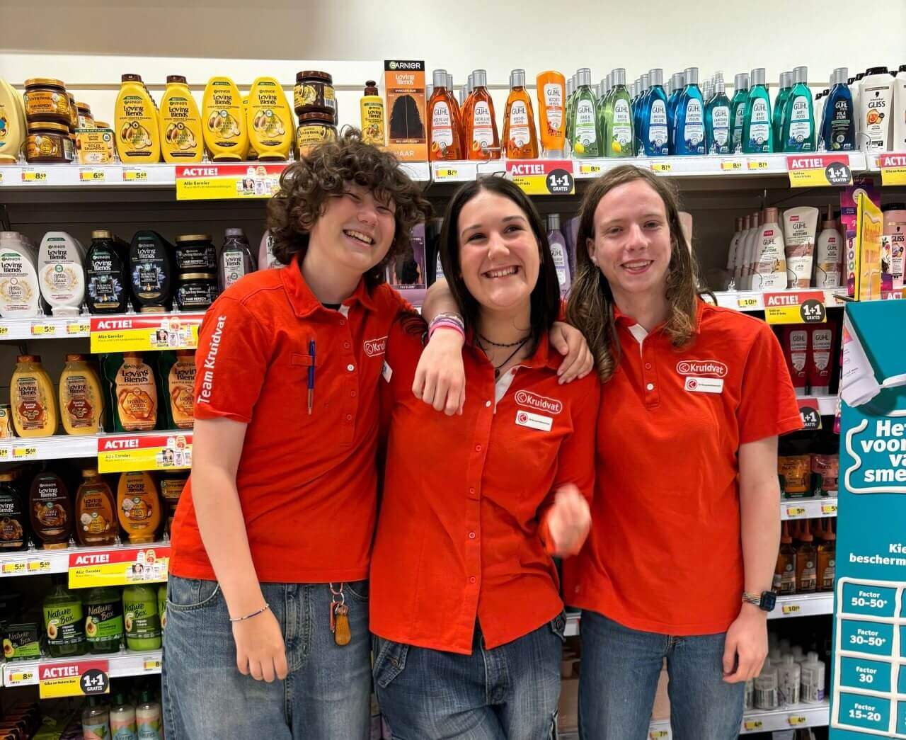 Three smiling teens in a Kruidvat drugstore, wearing red company shirts 