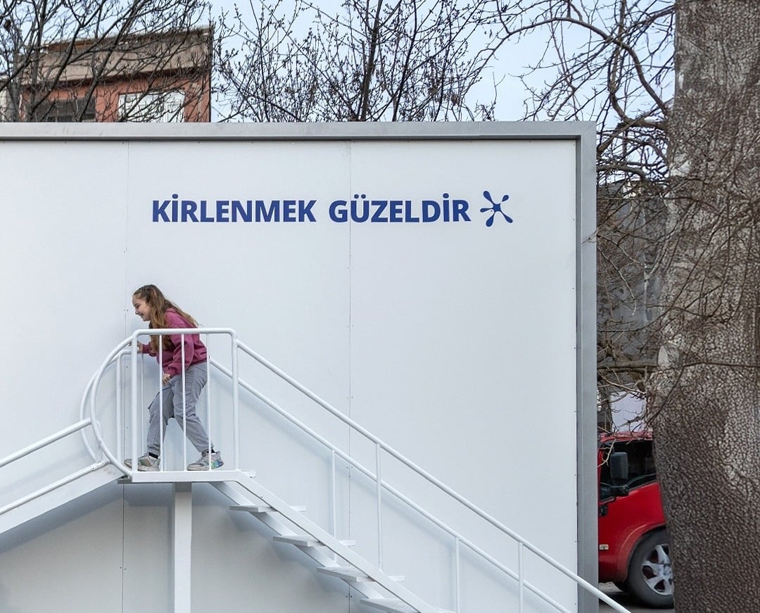 Girl about to go down a slide attached to a plain white billboard with the words 'Dirt is Good' in Turkish 