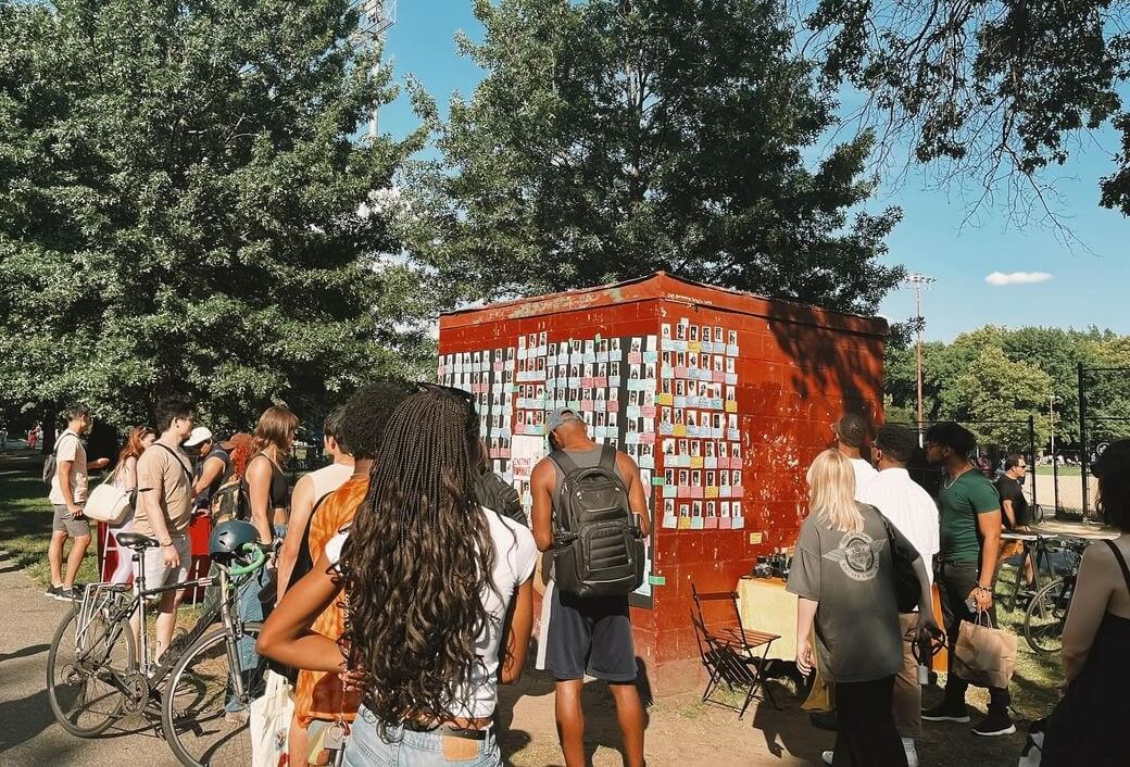 People gathering around the 'love wall' in McCarren Park, covered in photos of others 